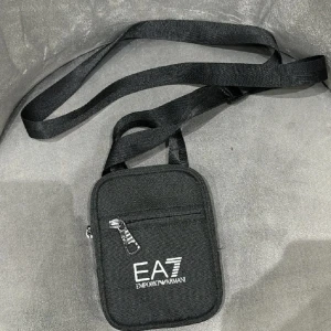 Svart axelväska från Emporio Armani - Snygg och praktisk svart axelväska från Emporio Armani med justerbar rem. Väskan har en dragkedja framtill och logotypen EA7 broderad i vitt. Perfekt för att bära med sig det viktigaste på ett stilfullt sätt.