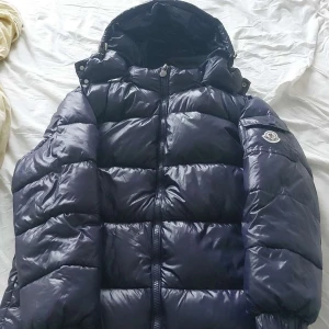 Svart dunjacka från Moncler - Säljer en svart, glansig dunjacka från Moncler med huva och dragkedja framtill. Jackan har en klassisk pufferstil och Moncler-logga på ärmen. Perfekt för kyliga dagar när du vill hålla dig varm och snygg.