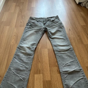 Grå jeansbyxor med broderi och dragkedjedetaljer - Säljer ett par grå jeansbyxor från Replay med snygga broderier och dragkedjor på bakfickorna. Byxorna har normal passform och raka ben. Perfekta för dig som vill ha något unikt i garderoben!