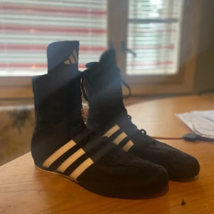 Svarta boxningsskor från Adidas med vita ränder - Säljer ett par svarta Adidas boxningsskor med klassiska vita ränder på sidorna och hög skaft. Skorna har snörning hela vägen upp och en platt, vit sula med bra grepp. Perfekta för träning eller match.