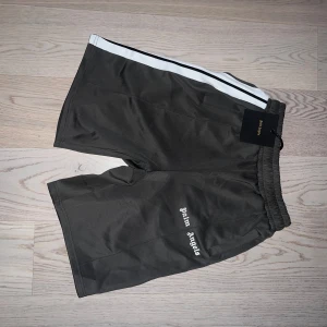 Grå shorts från Palm Angels med vit rand - Snygga grå shorts från Palm Angels med vit och svart rand längs sidorna. Broderad logga på ena benet och elastisk midja med snörning. Perfekta för en avslappnad stil.