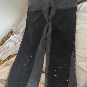 Gråsvarta jeansbyxor från Zara - Unika gråsvarta jeansbyxor från Zara med kontrasterande svarta paneler framtill och bakfickor. Byxorna har raka ben och detaljer med vita färgstänk på benen. Perfekta för dig som vill sticka ut med din stil.