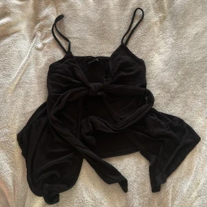 Svart knytlinne med smala axelband - Säljer ett svart linne med smala axelband och knytdetalj framtill. Toppen har en croppad passform och är perfekt för varma dagar. Snygg att styla med jeans eller kjol.