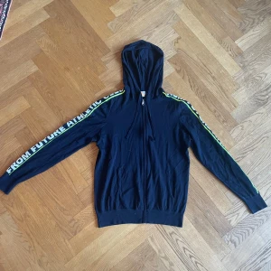 From Future zip hoodie  - Mörkblå hoodie med huva från From Future. Hoodien har dragkedja framtill och texten 'FROM FUTURE ATHLETIC CLUB' i vitt och grönt längs båda ärmarna. Tillverkad i 100% ull. NYPRIS 1500 / MITT PRIS 400!