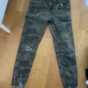 Cargopants i camouflage med slitningar - Säljer ett par tighta cargopants i camouflage-mönster med slitna detaljer och råa benslut. Byxorna har fickor framtill och är perfekta för en cool och avslappnad stil.