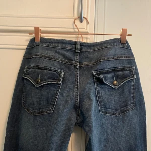 Blå bootcut jeans med ficklock - Snygga blå jeans med bootcut passform och ficklock bak med knappar. Klassisk femficksmodell och låg midja. Perfekta för dig som gillar en avslappnad men ändå trendig stil.