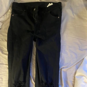 Svarta jeans med hål på knäna - Svarta jeans med raka ben och slitna hål på båda knäna. Klassisk femficksmodell med knapp och dragkedja i midjan. Perfekta för en avslappnad och trendig look.