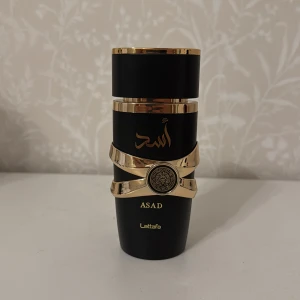 Asad Eau de Parfum från Lattafa 95-100ml - Säljer en lyxig parfymflaska från Lattafa, modell Asad. Flaskan är svart med eleganta gulddetaljer och arabiska inskriptioner. Den har en unik form och ett dekorativt guldband runt mitten. Perfekt för dig som gillar exklusiva dofter. Luktar som dior sauvage elixir