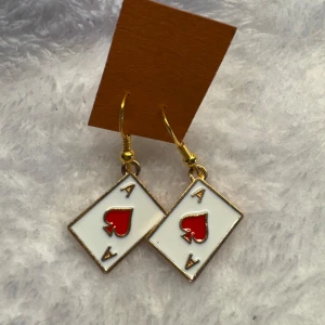 Örhängen med spelkortsmotiv - Unika örhängen i form av ess i hjärter, med vit bakgrund och röda hjärtan. Kanterna och krokarna är i guldfärg. Perfekt accessoar för dig som gillar spel eller vill sticka ut med något annorlunda. Krokarna är i sterling silver 