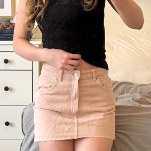 Ljusrosa jeanskjol - Säljer en ljusrosa kort jeanskjol med klassisk femficksdesign och knapp- samt dragkedjestängning framtill. Kjolen har bälteshällor och rå kant nertill för en avslappnad look. Perfekt för varma dagar och enkel att matcha.