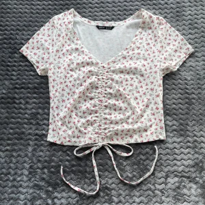 Vit blommig topp - Söt vit croppad topp med små rosa blommor och v-ringning. Toppen har korta ärmar och knytband framtill som ger en snygg rynkad effekt. Perfekt för varma dagar.