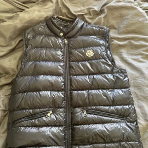 Svart dunväst från Moncler - Snygg svart dunväst från Moncler med klassisk logotyp på bröstet och dragkedja framtill. Västen har två snedställda fickor med dragkedja och quiltad design. Perfekt för lager-på-lager och stilren look.