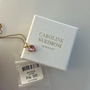 Halsband med hjärthänge från Caroline Svedbom - Säljer ett elegant halsband från Caroline Svedbom med ett hjärta i ljusrosa sten. Kedjan är i guldton och hänget har en glittrig finish. Perfekt för dig som gillar feminina och stilrena smycken. Har aldrig använt den!!!