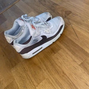 Nike Air Max 90 vita och svarta sneakers - Säljer ett par klassiska Nike Air Max 90 sneakers i vitt och svart med röda detaljer på tungan. Skorna har snörning, rund tå och den ikoniska Air Max-sulan för extra komfort. Perfekta för dig som gillar streetwear och sportig stil.