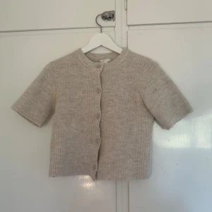 Trekvartsärmad kofta - Säljer en beige, med korta ärmar och rund hals. Koftan har knappar framtill och är croppad i modellen. Perfekt för lager-på-lager och enkel att matcha med olika outfits.