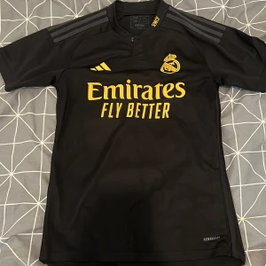 Real Madrid svart fotbollströja från Adidas - Säljer en svart Real Madrid fotbollströja från Adidas med gula detaljer och klubbmärke på bröstet. Tröjan har korta ärmar, rund hals och är tillverkad i 100% polyester. Perfekt för dig som vill visa ditt stöd för laget.