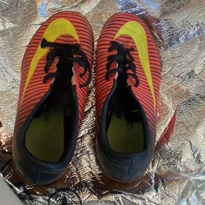 Nike fotbollsskor med röd och gul design - Säljer ett par Nike fotbollsskor med röd bas och svarta ränder samt en stor gul Nike-swoosh på sidan. Skorna har snörning och platt sula, perfekt för inomhusspel. Insidan är grön och yttersulan är brun.