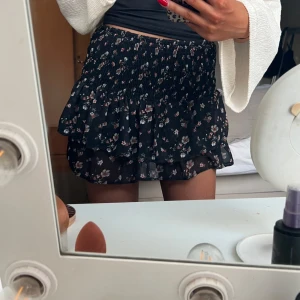 Svart blommig volangkjol - Supersöt svart kortkjol med småblommigt mönster och volanger. Kjolen har resår i midjan och är luftig med flera lager. 