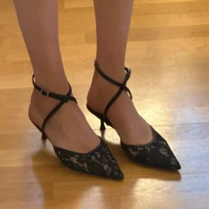 Svarta spetsiga kitten heels med spetsdetaljer - Eleganta svarta kittenheels med spetsig tå och fina spetsdetaljer. Skorna har en smal klack och justerbara remmar som korsas över foten för en stilren look. Aldrig använda, helt nya från NAKD, köpt för 599kr. Box finns kvar. Kompletterar en enkel outfit så snyggt och är väldigt bekväma!🤍