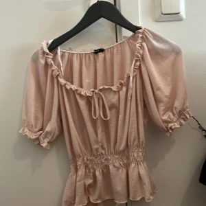Ljusrosa blus med volang och knyt - Söt ljusrosa blus med korta puffärmar, volangkant vid halsringningen och midjan samt knyt framtill. Blusen har resår i midjan och vid ärmsluten för en feminin siluett. Perfekt för vår och sommar! Den är använd en gång men var inte min stil 