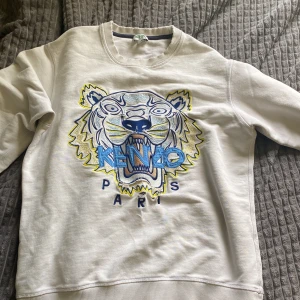 Vit sweatshirt med tiger från Kenzo - Säljer en vit sweatshirt från Kenzo med ikoniskt broderat tigermotiv och texten 'KENZO PARIS' i blått och gult framtill. Tröjan har rund halsringning och långa ärmar. Perfekt statement-plagg för dig som gillar streetwear.