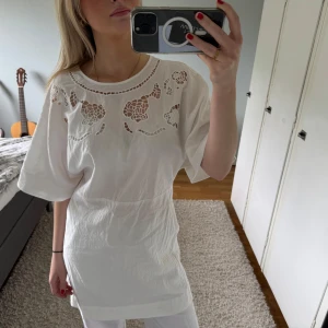 Vit klänning med broderade detaljer - Vit klänning med korta ärmar och unika broderade detaljer upptill. Blusen har en lös passform men insydd i midjan och rund halsringning. Den har två knytningar där bak. Perfekt för en stilren och luftig sommarlook.