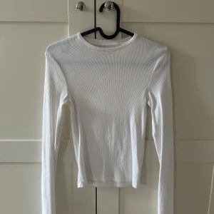 Vit ribbad långärmad topp - Säljer en enkel och stilren vit ribbad långärmad topp. Perfekt att matcha med jeans eller kjol för en clean look. Tunn och stretchig i materialet med rund halsringning. Aldrig använd!