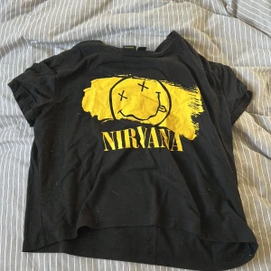 Svart Nirvana t-shirt med gul tryck - Svart t-shirt från Nirvana med ikoniskt gult smiley-tryck och text på framsidan. T-shirten har korta ärmar och en croppad passform. 