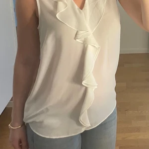 Vit blus med volang - Säljer en fin vit blus med volangdetalj framtill!💕Blusen är ärmlös och har en lätt v-ringning. Perfekt att matcha med jeans eller kjol för en fräsch look!💕