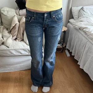 Blå bootcut jeans  - Säljer ett par mörk blå bootcut jeans med slitna detaljer. Jeansen har låg midja och bootcut stil. Storleken är w29 men passar mer som en w25 (s).