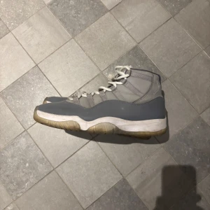 Gråa sneakers Nike Air Jordan 11 - Säljer ett par gråa Nike Air Jordans med högt skaft och vita snören. Skorna har en rund tå och platt sula, samt detaljer i olika nyanser av grått. Perfekta för dig som gillar klassiska sneakers med ikonisk design.