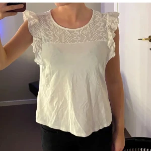 Vit blus med spets och volangärmar - Säljer en söt vit blus med spets upptill och volangdetaljer på ärmarna. Blusen är ärmlös och har rund halsringning. Perfekt för en fräsch och somrig look!