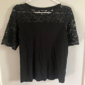 Svart blus med spetsdetaljer från Lindex - Säljer en svart blus från Lindex med vackra spetsdetaljer upptill och på ärmarna. Blusen har korta ärmar och rund halsringning. Perfekt för dig som vill ha en stilren men ändå elegant topp.