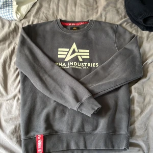 Grå sweatshirt från Alpha Industries - Säljer en grå sweatshirt från Alpha Industries med logga och tryck framtill. Tröjan har rund halsringning, långa ärmar och ribbade muddar. Klassisk och stilren modell med röd detalj nertill.