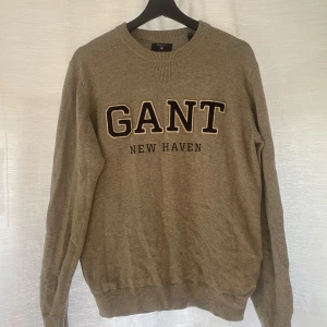Grå stickad tröja från GANT - Grå stickad tröja från GANT med broderad text 'GANT NEW HAVEN' på bröstet. Tröjan har rund halsringning, långa ärmar och ribbade muddar vid ärmslut och nederkant. Perfekt för en stilren och avslappnad look.