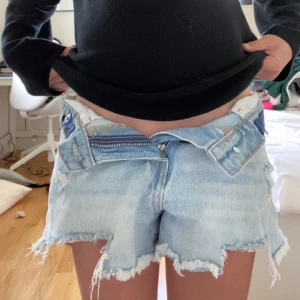 Ljusblå jeansshorts med slitningar - Säljer ett par ljusblå jeansshorts med råa kanter och slitna detaljer framtill. Shortsen har klassisk femficksdesign, gylf med dragkedja och knapp samt låg midja. Perfekta för varma dagar.