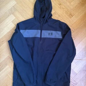 Svart vindjacka från Under Armour - Säljer en svart vindjacka med huva från Under Armour. Jackan har en grå panel över bröstet med logga och dragkedja framtill. Perfekt för blåsiga dagar och enkel att matcha med det mesta.