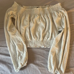 Vit offshoulder blus från HM - Säljer en söt vit offshoulder blus från HM med långa ärmar och smockdetaljer vid hals, midja och ärmslut. Blusen har ett diskret prickigt mönster i tyget och volangkant upptill. Perfekt för vår och sommar!