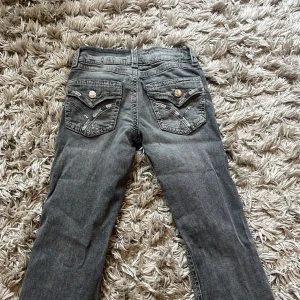 Grå bootcut jeans med dekorerade bakfickor - Säljer ett par grå jeans med bootcut passform. Byxorna har snygga dekorationer och knappar på bakfickorna som ger en unik look. Perfekta för dig som vill ha något extra till din outfit. Dem passar inte längre