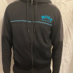 Svart hoodie från Hugo Boss med blå detaljer, (vi kan diskutera pris) - Säljer en svart hoodie från Hugo Boss med blå logga och linje över bröstet. Tröjan har dragkedja, huva med snörning och gråa paneler längs ärmarna. Perfekt för en sportig och avslappnad stil.