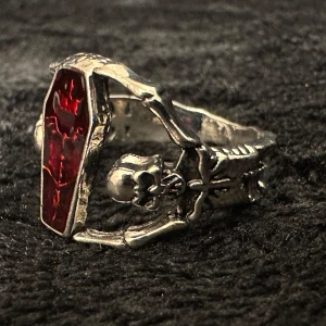 Silverfärgad ring med röd kista och dödskalle - Statementring i silverfärgad metall med kistformad front i djupröd resin och detaljerade dödskallar i bandet. Justerbar storlek. Innerdiameter ca 1,9 cm, höjd ca 1,5 cm. Nyskick, ej stämplad. Priset är fast – bundlerabatt vid köp av flera!  