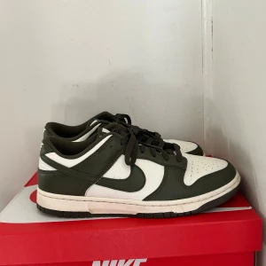 Nike Dunk Low i olivgrön och vit - Säljer ett par helt nya Nike Dunk Low med olivgröna och vita paneler. Använda en gång. Skorna har klassisk låg siluett, snörning och rund tå. Perfekta för dig som gillar streetwear och vill ha en stilren sneaker med snygga färgkontraster. 