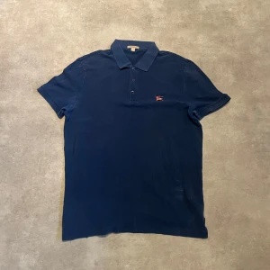 Marinblå pikétröja från Burberry - Säljer en klassisk marinblå pikétröja från Burberry med liten broderad logga på bröstet. Tröjan har krage och knäppning med tre knappar framtill. Helt ok skick, säljer billigt därav. Passar både L och M