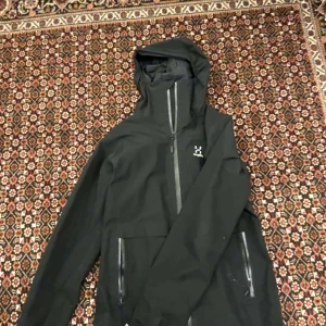Haglöfs jacka - Säljer en svart skaljacka från Haglöfs med huva och dragkedjor framtill samt vid fickorna. Jackan har en minimalistisk design och är perfekt för utomhusaktiviteter. Märkeslogga på bröstet och insidan.