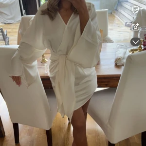 Vit omlottklänning med knytband (student) - Säljer en vit omlottklänning med vida ärmar och knytband i midjan. Klänningen har en kort längd och är tillverkad i ett glansigt material som ger en lyxig känsla. Perfekt för sommarens tillfällen när du vill känna dig extra fin. Passar även bra till student, använd en gång