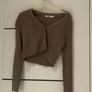 Beige ribbad kofta från NA-KD - Säljer en beige ribbad kofta från NA-KD i storlek M. Koftan är croppad med v-ringning och knappar framtill. Perfekt för lager-på-lager och har långa ärmar. Snygg och trendig modell! Aldrig använd! 