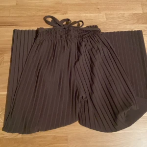 Svarta plisserade wide leg byxor - Säljer ett par svarta plisserade byxor med breda ben och resår i midjan. Byxorna har en flowy känsla och knytband i midjan för extra detalj. Perfekta för en trendig och avslappnad stil.