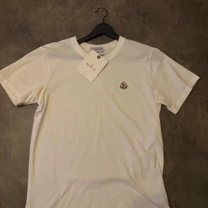 Vit t-shirt från Moncler - Säljer en stilren vit t-shirt från Moncler med klassisk rund hals och Moncler-logga broderad på bröstet. T-shirten är enkel och passar perfekt till en avslappnad look. Materialet känns mjukt och skönt mot huden. Storlek L men passar även M