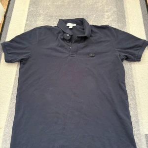 Mörkblå Lacoste pikétröja slim fit - Snygg mörkblå pikétröja från Lacoste i slim fit-modell. Klassisk krage med två knappar och diskret broderad krokodil på bröstet. Tillverkad i mjuk bomull som känns skön mot huden. Perfekt för en clean och sportig look.
