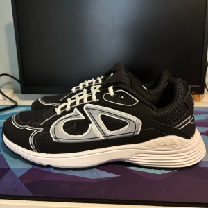 Svarta Dior B30 sneakers med vita detaljer - Snygga Dior B30 sneakers i svart med vita och grå detaljer. Skorna har en chunky vit sula, meshpaneler och stora CD-loggor på sidorna. Klassisk snörning och sportig vibe, perfekt för dig som gillar exklusiva streetwear-inspirerade sneakers.